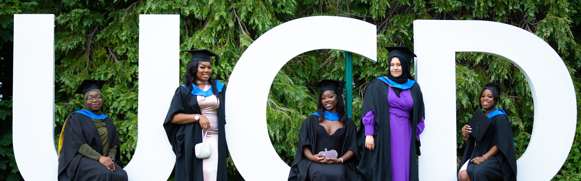 Five UCD alumnae posing by UCD letters at their graduation.